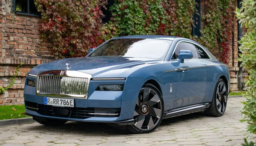 Niebieski Rolls-Royce Spectre, luksusowy samochód elektryczny, zaparkowany przed ceglanym budynkiem porośniętym jesiennymi liśćmi.