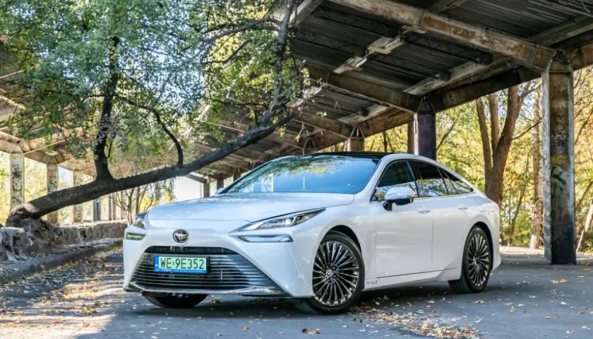 Biała Toyota Mirai, samochód przyszłości, zaparkowana pod zadaszeniem z opadłymi liśćmi wokół.