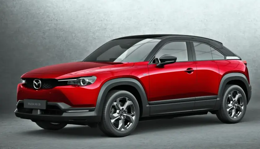 Czerwona Mazda MX-30 z czarnym dachem, stojąca na tle szarej ściany. Stylowy crossover miejski.