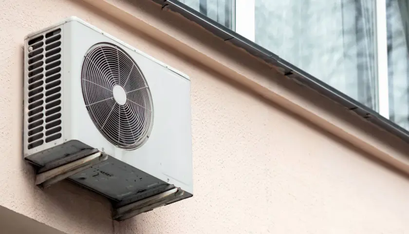 Klimatyzacja w bloku, biała jednostka zewnętrzna zamontowana na jasnoróżowej ścianie, z widocznym wentylatorem i kratką.