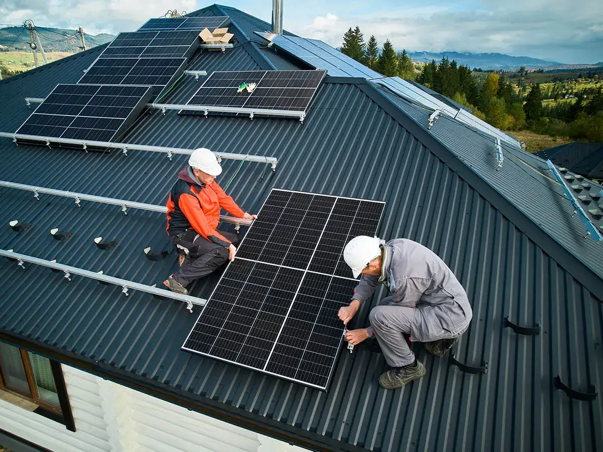 Dwóch pracowników w kaskach montuje panele fotowoltaiczne na dachu. Praca nad zieloną energią w malowniczej okolicy.