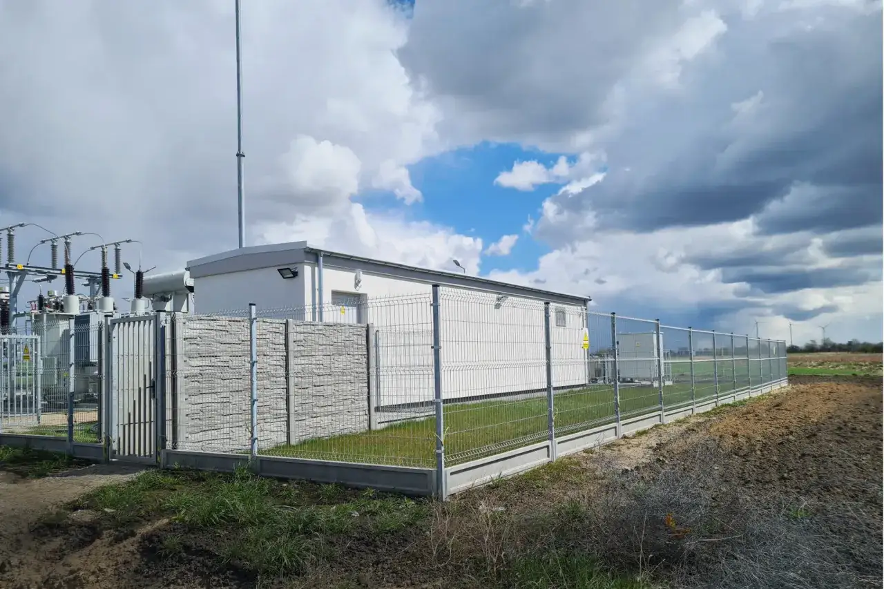 Nowoczesna farma fotowoltaiczna z transformatorami i budynkiem technicznym, otoczona ogrodzeniem.